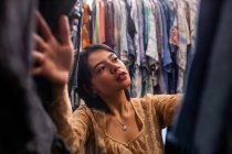 Attractive young lady searching for new outfit on clothes rail in small shop — Stock Photo