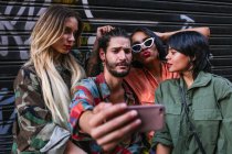 Group of cheerful young people in trendy outfits standing on street of modern city and taking selfie together — Stock Photo