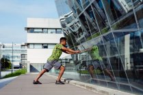 Seitenansicht eines erwachsenen ethnischen Mannes, der sich am gläsernen Geländer dehnt, während er auf der modernen Stadtstraße trainiert — Stockfoto