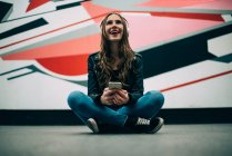 Happy young woman sitting on floor with mobile phone and listening to music against colorful background — Stock Photo