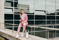 Fashionable young woman in pink suit with coffee and phone sitting on railing in front of modern office building — Stock Photo