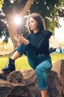 Jovem mulher sentada no rock e segurando smartphone no parque — Fotografia de Stock