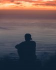 Back view of unrecognizable person sitting on rock and admiring sunset at the ocean. - foto de stock