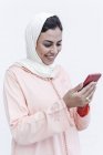 Mujer marroquí sonriente con hijab y vestido árabe típico usando el teléfono móvil sobre fondo blanco - foto de stock