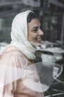 Sonriente mujer marroquí con hijab sentado detrás del cristal de la ventana - foto de stock
