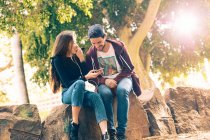 Riendo joven pareja sentado en roca con teléfono inteligente en el parque - foto de stock