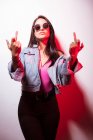 Contemptuous young pink dressed woman in sunglasses standing at white wall and showing middle fingers — Stock Photo
