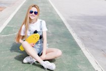Blonde fille élégante assise sur skate park avec penny board et regardant la caméra — Photo de stock