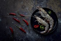 Сырые тигровые креветки с маленьким красным перцем на тарелке на темной столешнице — стоковое фото