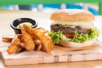 Appetizing unhealthy burger served with French fries and sauce on cutting board — Stock Photo