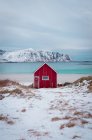 Petite cabane en bois rouge sur le littoral enneigé avec eau de mer bleue et montagnes en arrière-plan, Lofoten, Norvège — Photo de stock