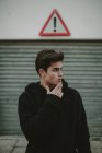 Thoughtful teenager in black hooded jacket standing on street with exclamation sign and looking away — Stock Photo
