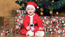Portrait of happy little boy in Santa Claus costume sitting at Christmas tree — Stock Photo