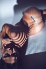 Nackte junge brünette Frau mit Tätowierung auf dem Rücken und gestreiftem Schatten auf Gesicht und Körper im Studio liegend — Stockfoto
