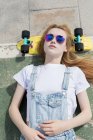 Blonde girl in sunglasses lying on footpath on penny board — Stock Photo