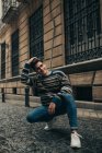 Young smiling stylish teenager in sweater crouching on city street and looking at camera — Stock Photo
