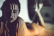 Mujer joven seductora desnuda tumbada en el estudio con sombra rayada en la cara y el cuerpo - foto de stock
