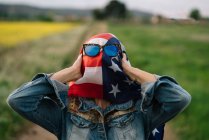 Signora in abiti casual soggiorno in campo giallo con occhiali da sole sopra la bandiera americana con la luce del sole — Foto stock