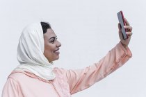 Femme marocaine souriante avec hijab et robe arabe typique prenant selfie sur fond blanc — Photo de stock