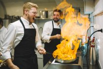 Cocinero feliz haciendo un flambe en la cocina del restaurante con colega viendo - foto de stock