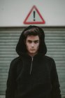 Adolescent réfléchi en veste à capuche noire debout sur la rue avec signe d'exclamation et en regardant la caméra — Photo de stock