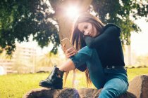 Giovane donna sorridente seduta sulla roccia e utilizzando smartphone nel parco — Foto stock