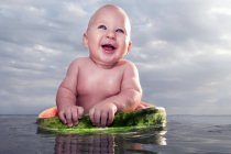 Cheerful nude toddler boy sitting in watermelon on water — Stock Photo