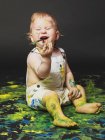 Adorable dirty little boy sitting and playing with yellow and blue paint on dark background. — Stock Photo