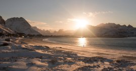 Costa nevada con playa de arena y mar a la luz del sol en el fondo de las montañas, Lofoten, Noruega - foto de stock