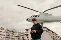 Bonito jovem de pé no monumento de helicóptero na cidade e usando smartphone — Fotografia de Stock