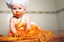 Cheerful little child sitting on tale and having fun while eating pasta from bowl. — Stock Photo