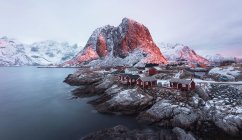Pittoresca vista del piccolo villaggio con case in legno rosso sulla costa rocciosa del porto calmo mare con montagne, Lofoten, Norvegia — Foto stock