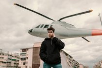 Handsome young man standing at helicopter monument in city and talking on smartphone — Stock Photo