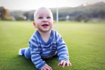 Young boy sitting on green lawn at the hole in golf club and looking at camera. — Stock Photo