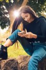 Молода усміхнена жінка сидить на камені і використовує смартфон у парку — стокове фото