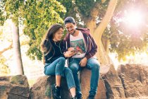 Laughing young couple sitting on rock with smartphone in park — Stock Photo