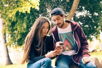 Joven pareja sentado y compartir smartphone en el parque - foto de stock