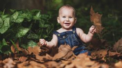 Cute little toddler in hat and denim clothes sitting and playing with foliage in nature. — Stock Photo