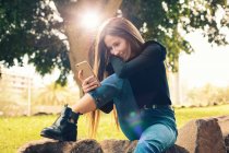 Молодая улыбающаяся женщина сидит на камне и использует смартфон в парке — стоковое фото