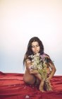 Jeune femme assise sur un drap rouge en lingerie et tenant des fleurs — Photo de stock