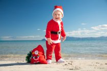 Cheerful little boy in Santa Claus costume standing on beach in sunny day — Stock Photo