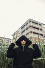 Uomo con cappuccio nero giacca in piedi nel cespuglio verde in città — Foto stock