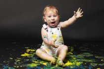 Liebenswerter schmutziger kleiner Junge sitzt und spielt mit gelber und blauer Farbe auf dunklem Hintergrund. — Stockfoto