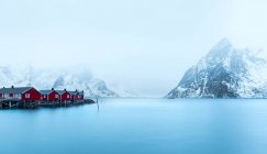 Landscape of small red wooden cabins on coast against snowy mountains in haze, Norway — Stock Photo