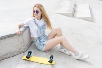 Blonde girl sitting sitting on asphalt with penny board — Stock Photo