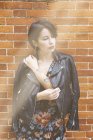 Вдумчивая татуированная женщина в узорчатом платье и кожаной куртке, стоящая перед кирпичной стеной — стоковое фото