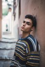 Молодой уверенный подросток в свитере, опирающийся на стену на городской улице и смотрящий в камеру — стоковое фото