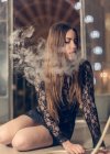 Симпатичная брюнетка в кружевном платье курит кальян чувственно сидя на полу с закрытыми глазами — стоковое фото