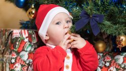 Petit garçon en costume de Père Noël assis à l'arbre de Noël et boule mordante — Photo de stock