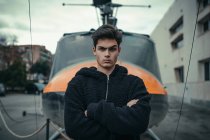 Young doubtful man standing with arms crossed with helicopter monument on background — Stock Photo